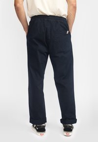 Man die donkerblauwe losse broek met elastische tailleband en opgerolde pijpen draagt, wit t-shirt en zwart-witte sneakers, staand met handen in de zakken.