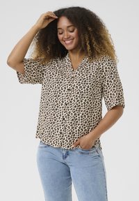 Blouse à manches courtes, imprimé léopard en beige et noir ; possède un col, des boutons sur le devant et une coupe décontractée. Portée avec un jean bleu clair.