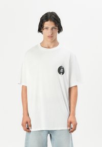 Weißes Baumwoll-T-Shirt mit rundem Halsausschnitt und kurzen Ärmeln, mit einem schwarzen kreisförmigen Grafiken auf der linken Brustseite.