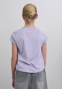 T-shirt in cotone viola chiaro con maniche corte, scollo rotondo e vestibilità comoda, abbinata a una gonna di jeans grigia. Design semplice, senza motivi visibili.
