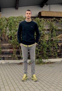 Abbigliamento informale con un maglione blu navy, una camicia a collare gialla, pantaloni grigio chiaro e sneakers gialle brillanti, ambientato tra verde e trame.