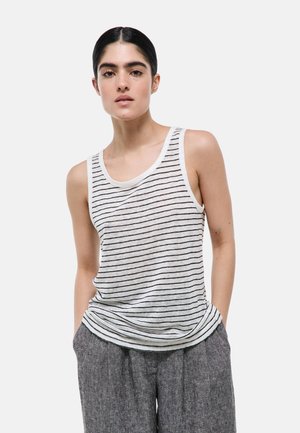 Femme aux cheveux foncés en chignon bas portant un débardeur blanc avec de fines rayures horizontales noires et un pantalon gris, mains dans les poches.