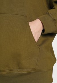 Mano descansando dentro del bolsillo frontal de un sudadera de manga larga verde olivo con puños acanalados y costura visible.