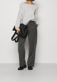 Vrouw in een grijze off-shoulder trui, losse donkergrijze broek, zwarte chunky schoenen, met in één hand een zwarte gewatteerde rugtas.