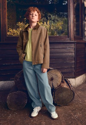 Enfant roux debout les mains dans les poches, portant une veste marron, une chemise vert clair, un jean bleu et des baskets blanches près d'un tas de bois empilé.