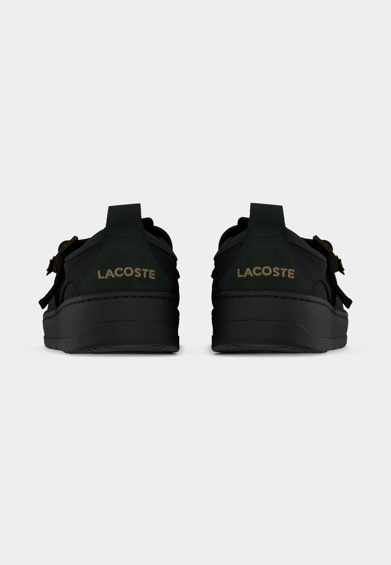 Scarpa da ginnastica nera con dettagli verdi, materiale strutturato e suola spessa. Presenta il nome del marchio "LACOSTE" in oro sul tallone.