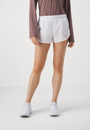 Kvinde iført hvide atletikshorts med grå Nike-logoer, hvide sneakers og en brun langærmet top, stående mod en ensfarvet baggrund.