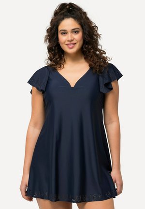 Woman with curly hair wearing a navy blue dress with flutter sleeves and laser-cut floral patterns at sleeves and hem, smiling at the camera.