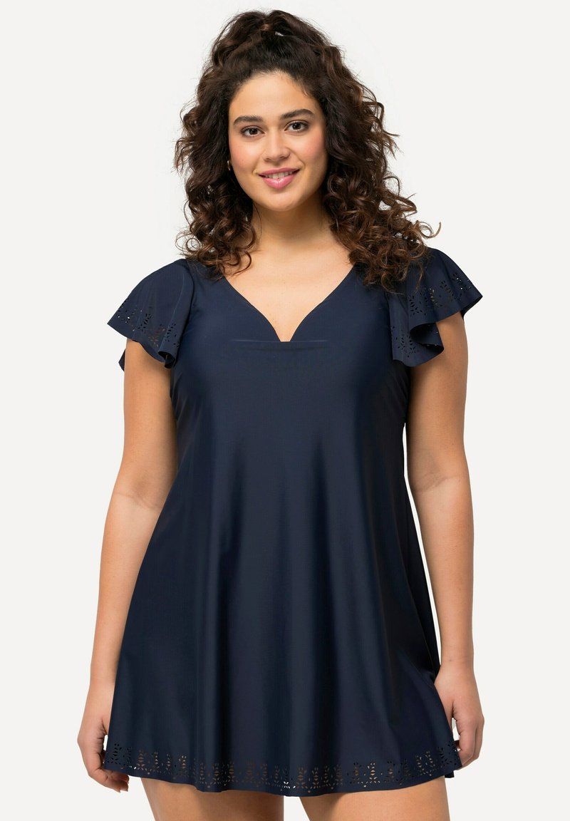 Donna con capelli ricci che indossa un abito blu navy con maniche svolazzanti e motivi floreali tagliati al laser su maniche e orlo, sorridente verso la fotocamera.