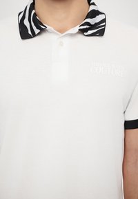 Camisa polo branca com um colarinho às riscas de zebra em preto e branco. Apresenta uma textura subtil, uma pala de botões dupla e um logótipo impresso no peito.