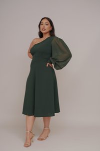 Robe verte à une épaule avec une manche ballon, corsage ajusté et jupe évasée. Associée à des talons dorés à lanières. Texture lisse.