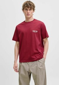 Burgundy bomulds t-shirt med korte ærmer og rund hals. Hvid trykt tekst på venstre bryst. Sammen med khaki bukser.