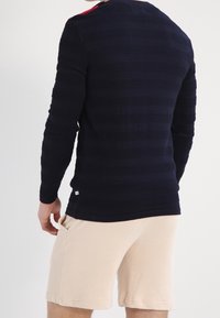 Pull en maille texturée bleu marine à manches longues avec des rayures horizontales, présentant un ourlet incurvé et un accent rouge centré dans le dos. Associé à un short beige.