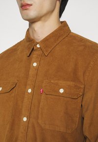 Homme portant une chemise en velours côtelé marron à boutons avec deux poches poitrine à boutons et une petite étiquette rouge sur la poche gauche.