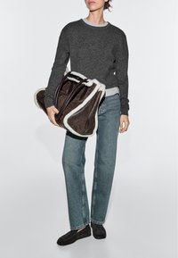 Person wearing dark gray sweater, blue jeans, and black loafers holding large brown bag with white shearling trim.