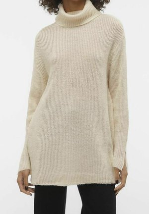 Femme portant un pull en maille crème à col roulé manches longues avec ouvertures pour les pouces, associé à un pantalon noir, sur un fond uni.