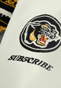 Biely kožený povrch s čiernym a žltým vyšitým logom pantera a slovom "SUB5CRIBE" v tučnej čiernej niťi.