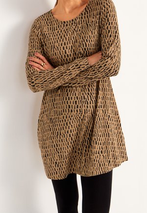 Kvinde iført en løstsiddende, langærmet beige tunika med sort stregmønster, armene krydsede, kombineret med sorte leggings.