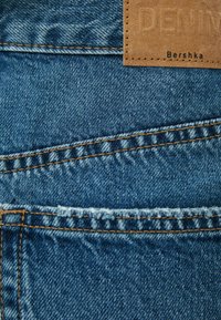 Jasnoniebieska tkanina dżinsowa z przeszyciami, z brązową skórzaną metką z tłoczeniem "DENIM" oraz "Bershka" dla identyfikacji marki.
