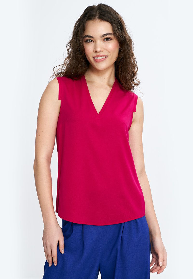 Fuksjowa, bezrękawnikowa bluzka z dekoltem w kształcie litery V i luźnym krojem, wykonana z gładkiego materiału. W zestawie z niebieskimi spodniami o szerokich nogawkach.
