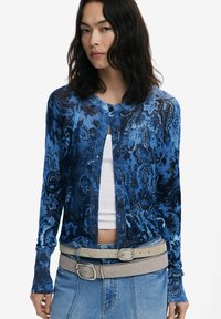 Cardigan bleu à motifs avec un col rond, des manches longues et une fermeture à boutons. Porté sur un haut blanc et associé à une ceinture grise et une jupe en denim.