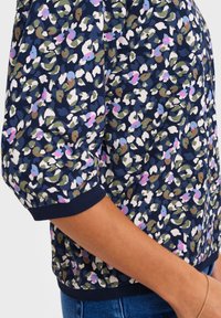 Frau, die eine marineblaue Bluse mit mehrfarbigem abstraktem Blumenmuster, dreiviertellangen Puffärmeln sowie dunkelblauen gerippten Bündchen und Saum trägt.