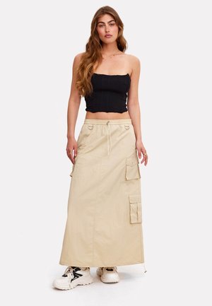 Femme aux longs cheveux portant un crop top noir sans bretelles, une jupe maxi cargo beige et des baskets blanches épaisses sur un fond uni.