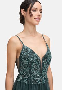 Zielona sukienka w odcieniu szmaragdowym z delikatnym beadingiem i kwiatowymi wzorami, prześwitującym środkowym panelem oraz regulowanymi ramiączkami, z warstwową spódnicą z tiulu.