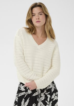 Young woman with shoulder-length blonde hair wearing a white knitted V-neck sweater and a black skirt with white leaf patterns.