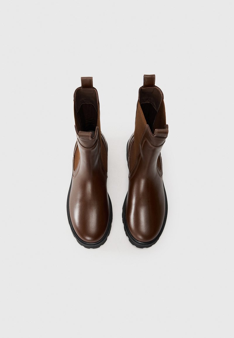 Ein Paar braune Leder-Chelsea-Stiefel mit schwarzen Sohlen und seitlichen elastischen Einsätzen, von oben auf einem schlichten weißen Hintergrund betrachtet.