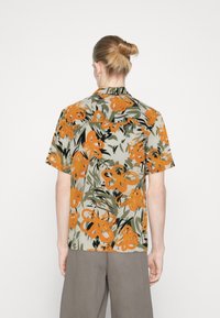 Person med lyst hår i en knold iført en kortærmet skjorte med et orange og grønt blomstermønster, stående mod en ensfarvet baggrund.