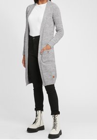 Long grey cardigan made of textured knit, featuring open front, side pockets, and ribbed cuffs. Worn over a white shirt and black jeans.