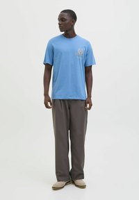 Tricou albastru din bumbac cu un design imprimat pe piept, asociat cu pantaloni largi de culoare maro închis și pantofi slip-on bej.