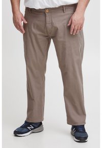 Muž v khaki chinos nohaviciach a námorníckych modrých teniskách New Balance stojí pred jednoduchým pozadím.