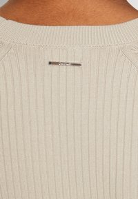 Beige knitted sweater featuring a ribbed texture and a silver logo tag at the back neckline, providing detail and subtle contrast.