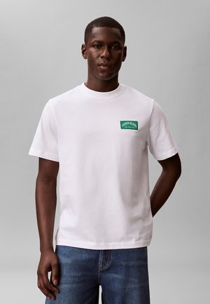 Mężczyzna w białej koszulce Calvin Klein z zieloną naszywką z logo na piersi i niebieskich dżinsach, stojący na jednolitym szarym tle.