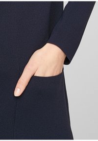 Granatowy materiał z prążkami z przednią kieszenią, z ręką częściowo wsuniętą w jej wnętrze. Gładka faktura, długie rękawy, dopasowany krój.