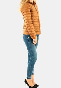 Doudoune marron avec piqûres horizontales, fermeture éclair et capuche. Portée sur un jean bleu ajusté et des talons noirs, mettant en valeur une silhouette élégante.