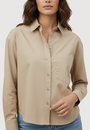 Femme portant une chemise beige à boutons avec une poche poitrine et un jean bleu, touchant l'ourlet de la chemise avec sa main droite.