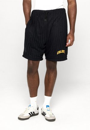 Hombre vistiendo pantalones cortos negros de malla con texto amarillo "COLUC", calcetas blancas y zapatillas Adidas blancas con rayas negras, de pie con las manos en los bolsillos.