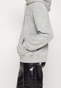 Felpa grigia con cappuccio e tasca frontale, dotata di un cordino per il cappuccio e una leggera texture. Abbinata a pantaloni neri lucidi con motivo coccodrillo.