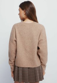 Maglione a maglia marrone chiaro con orlo a coste e maniche larghe. Abbinato a una gonna a scacchi. La texture sembra morbida e accogliente.