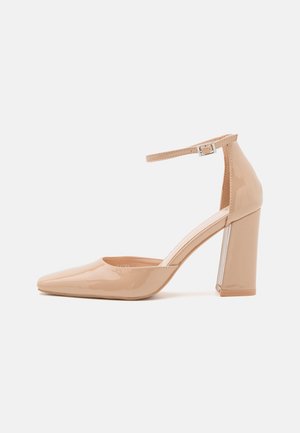 Beige gepatenteerd leren hoge hak schoen met een spitse neus, enkelsluiting met een gesp, en een chunky hak met een transparante accent.