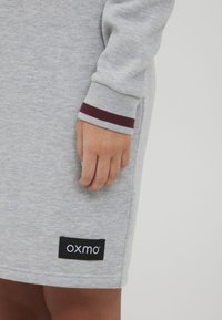 Siva haljina s duksericom s bordo prugastim rukavima. Ima crnu oznaku s bijelim текстом "oxmo" blizu rubа. Mekana tekstura tkanine.