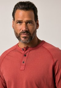 Rotes Langarm-Henley-Shirt aus weichem Stoff, mit einem Knopfverschluss und schwarzen Knöpfen sowie gestickten Akzenten an den Ärmel.