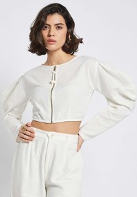 Weißer, verkürzter Blazer mit rundem Halsausschnitt und goldener Reißverschluss, ausgestattet mit Puffärmeln und strukturiertem Stoff. Kombiniert mit hochgeschnittenen Hosen.