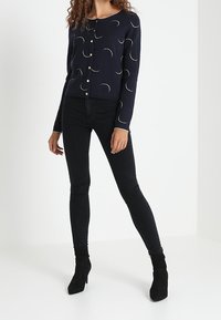 Granatowy sweter rozpinany z motywami półksiężyców, z guzikami z przodu, noszony na czarne dopasowane jeansy i botki za kostkę. Prosty, dopasowany krój.