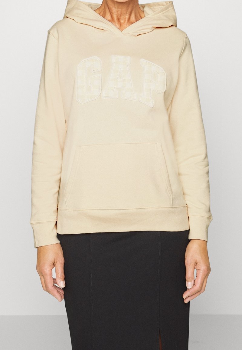 Beige hoodie met een voorzak en wit "GAP" logo. Zachte stof, lange mouwen en een capuchon met trekkoord. Draagt over een zwarte rok.