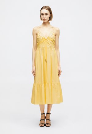 Moteris su trumpais plaukais, dėvinti geltoną midi suknelę be rankovių, su raukšlėtu liemeniu ir juodus aukštakulnius su dirželiais, stovinti ant paprasto balto fono.