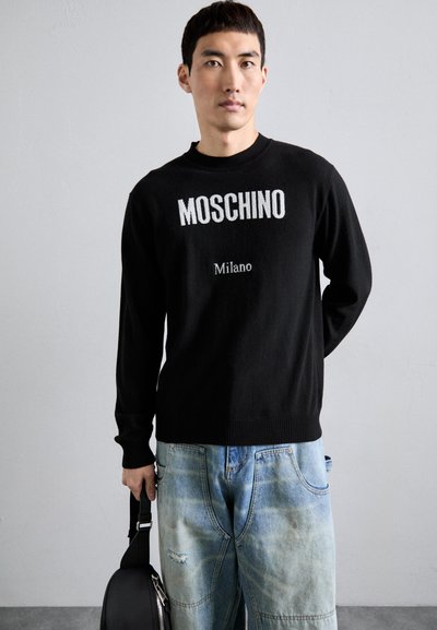Schwarzer Strickpullover mit "Moschino" und "Milano" in weißer Schrift, kombiniert mit hellblauen, ausgebeulten Jeans und einer schwarzen Schultertasche.